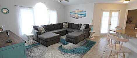 A living room with a sectional sofa, a ceiling fan, and a kitchen area with a bar stool.