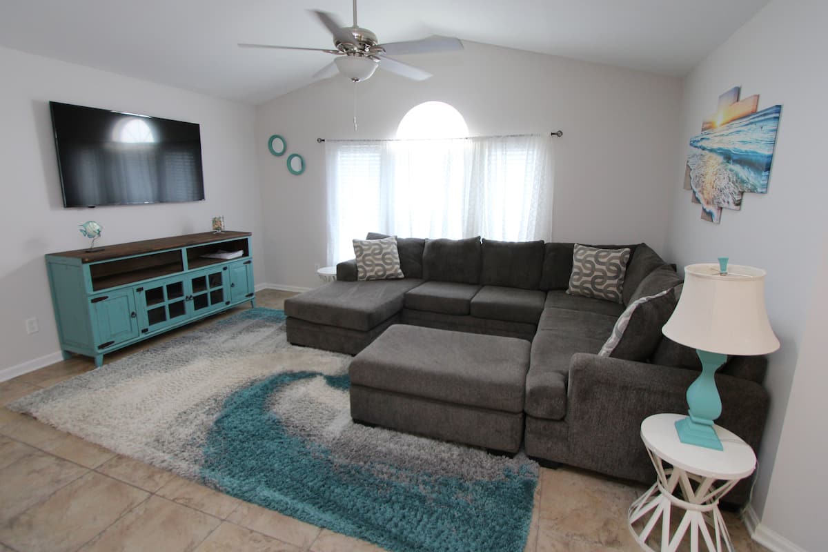 A living room with a sectional sofa, a teal entertainment unit, a ceiling fan, and a large TV on the wall.