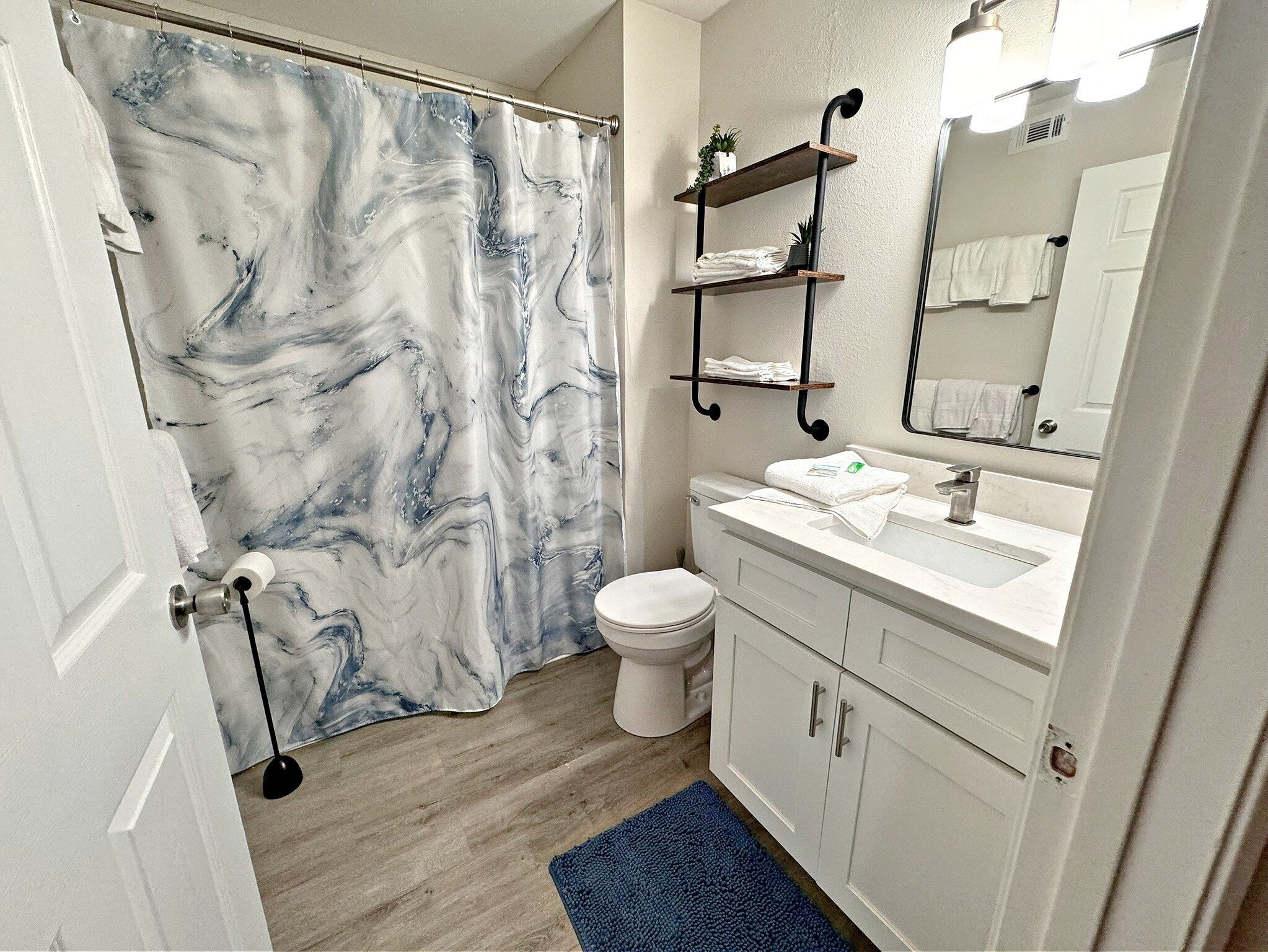 Een badkamer met een marmeren douchegordijn, een toilet, een wastafel met spiegel en een blauw vloerkleed.