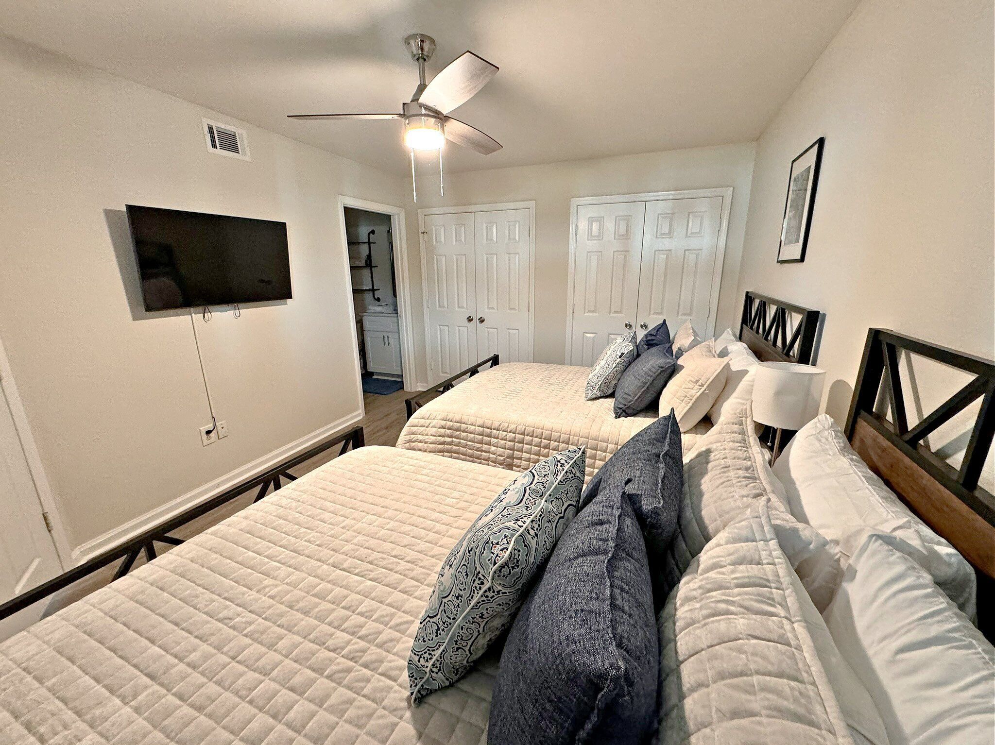 A bedroom with two beds, a ceiling fan, a TV mounted on the wall, and a door leading to a bathroom.