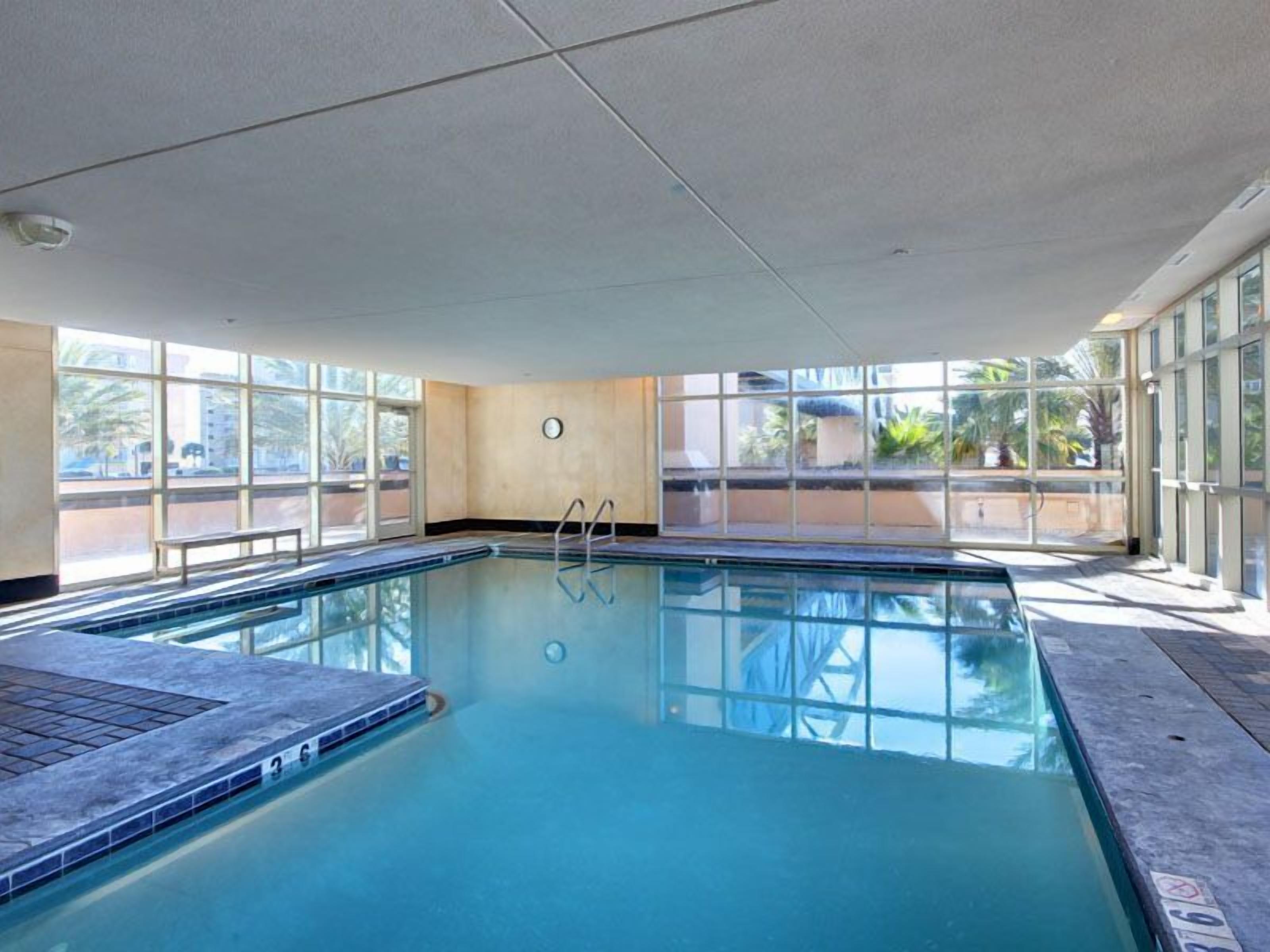 A large indoor swimming pool with clear glass walls, a ladder, and a view of the outside.