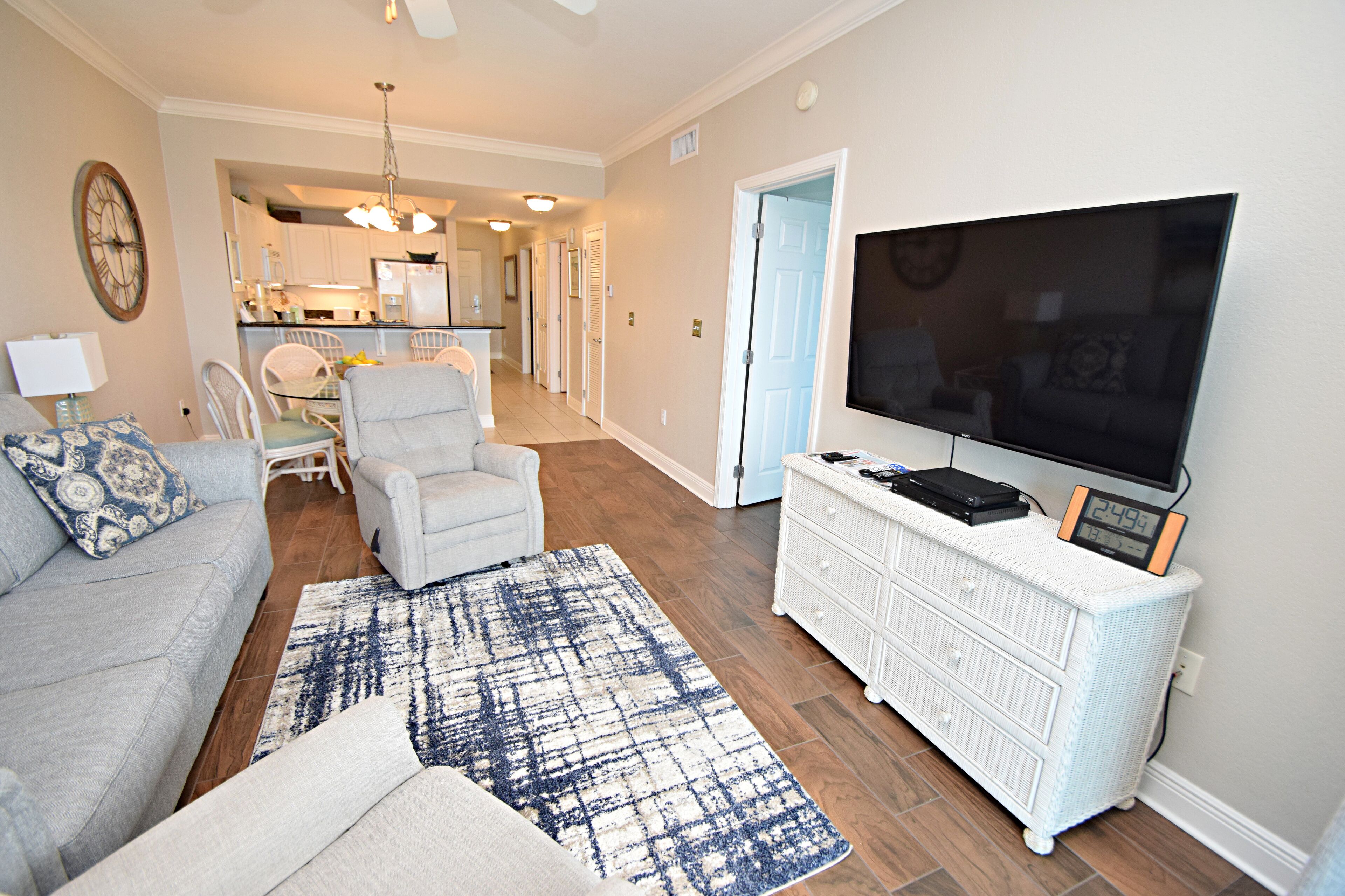 A living room with a grey sofa, a white armchair, a blue and white rug, a white dresser with a TV and clock, and a kitchen in the background.