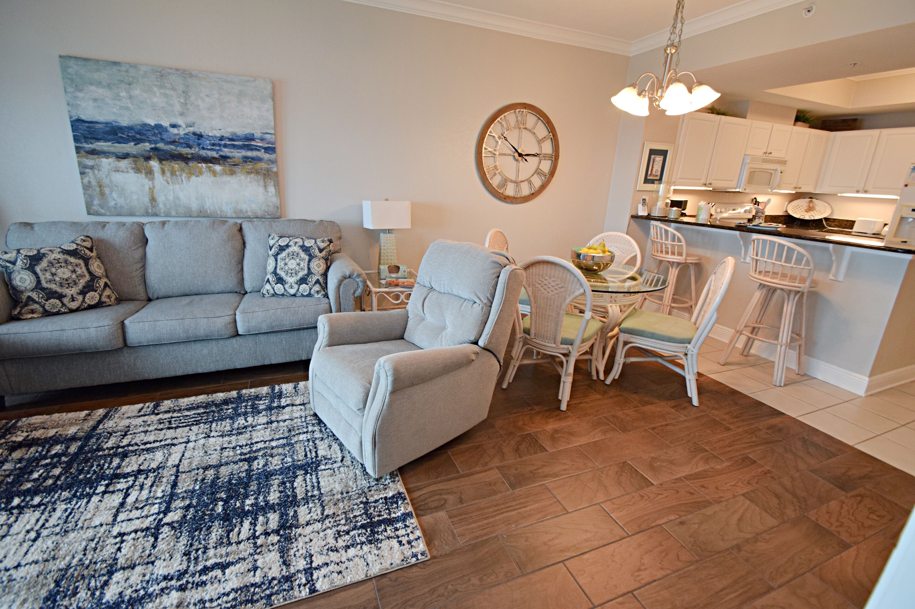 Un salon avec un canapé gris, des coussins à motifs, un tapis bleu et blanc, un fauteuil gris et un coin repas avec des chaises et une table blanches.