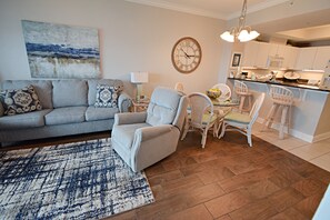 A living room with a grey sofa, patterned cushions, a blue and white rug, a grey armchair, and a dining area with white chairs and a table.