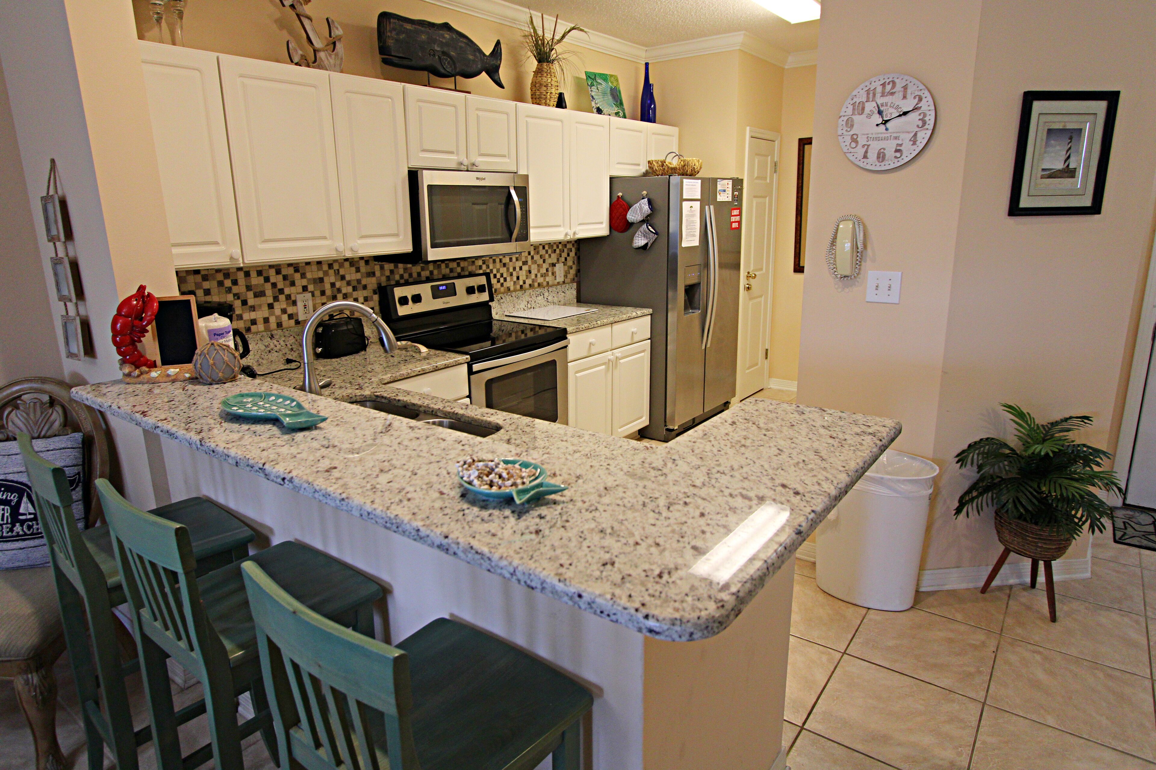 A kitchen with white cabinets, a granite countertop, a stainless steel refrigerator, a microwave, and a sink. There are two bar stools and a potted plant.
