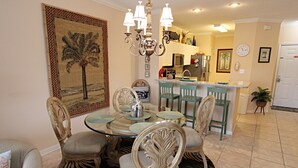 Uma sala de jantar com mesa de vidro, cadeiras e um candelabro. Dá para ver uma cozinha ao fundo, com armários e eletrodomésticos.