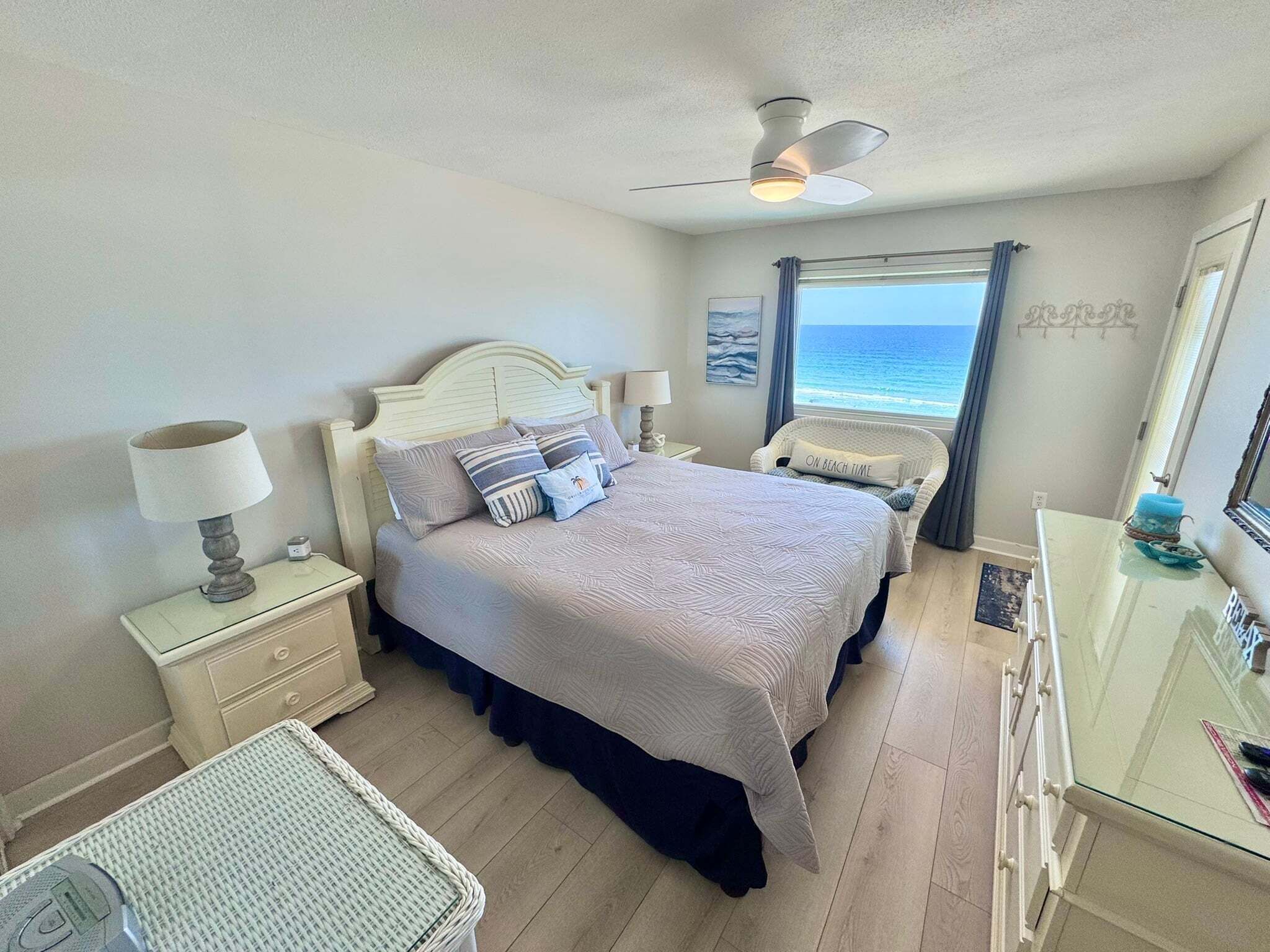 A bedroom with a bed, bedside tables, a chair, a dresser, and a view of the ocean.