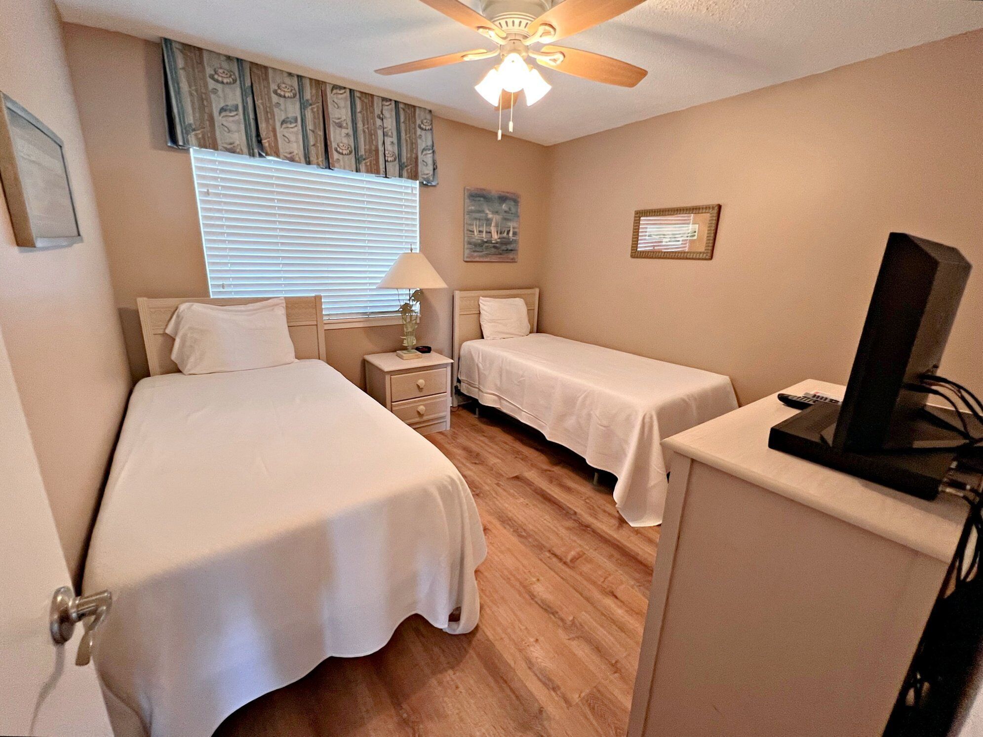 A hotel room with two beds, a ceiling fan, a TV, and a window with blinds.