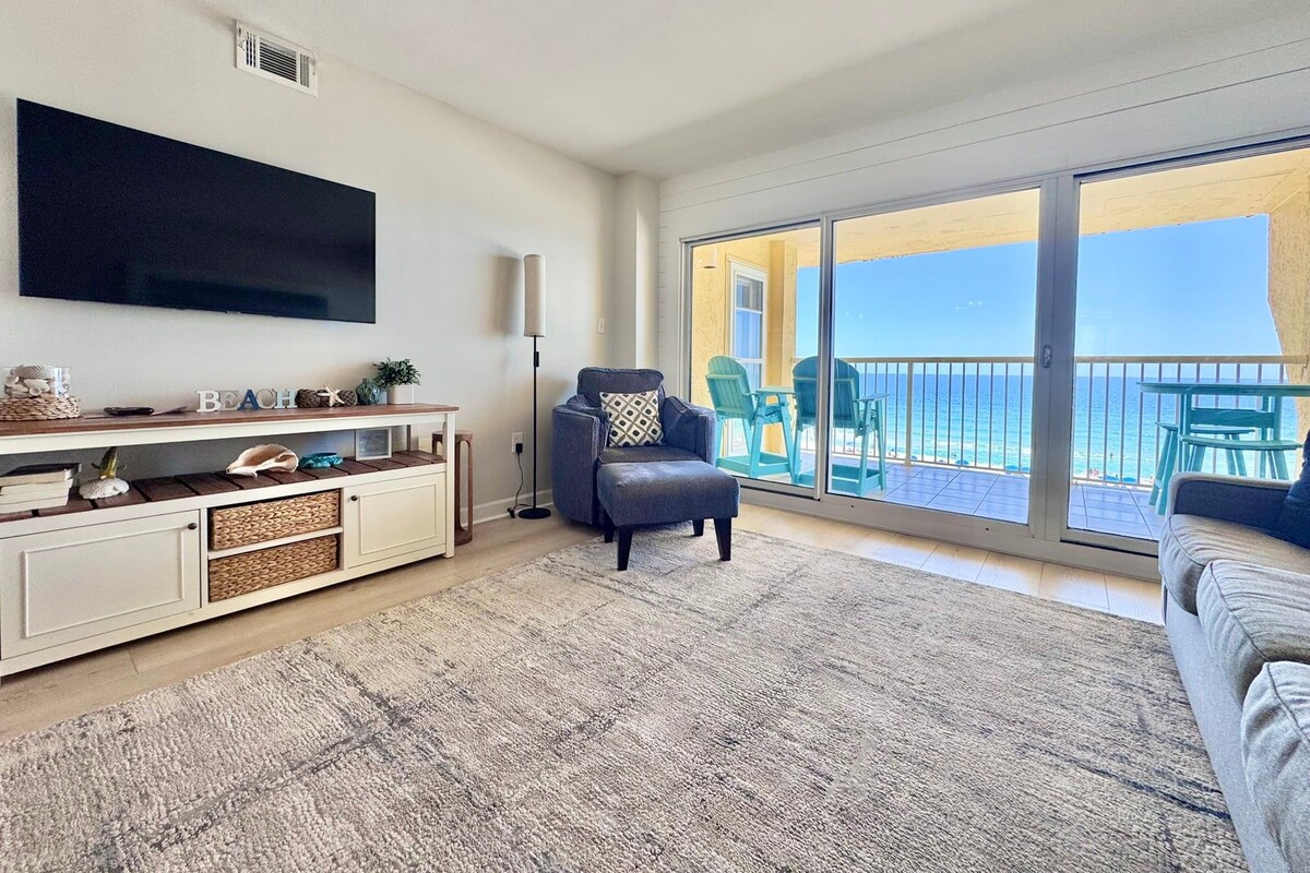 A living room with a flat-screen TV, a sofa, a chair, a small table, and a large window with a view of the ocean.