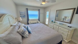 A bedroom with a bed, a dresser, a ceiling fan, a window with a view of the ocean, and a beach-themed pillow. - Driftwood Towers (Gulf Shores)