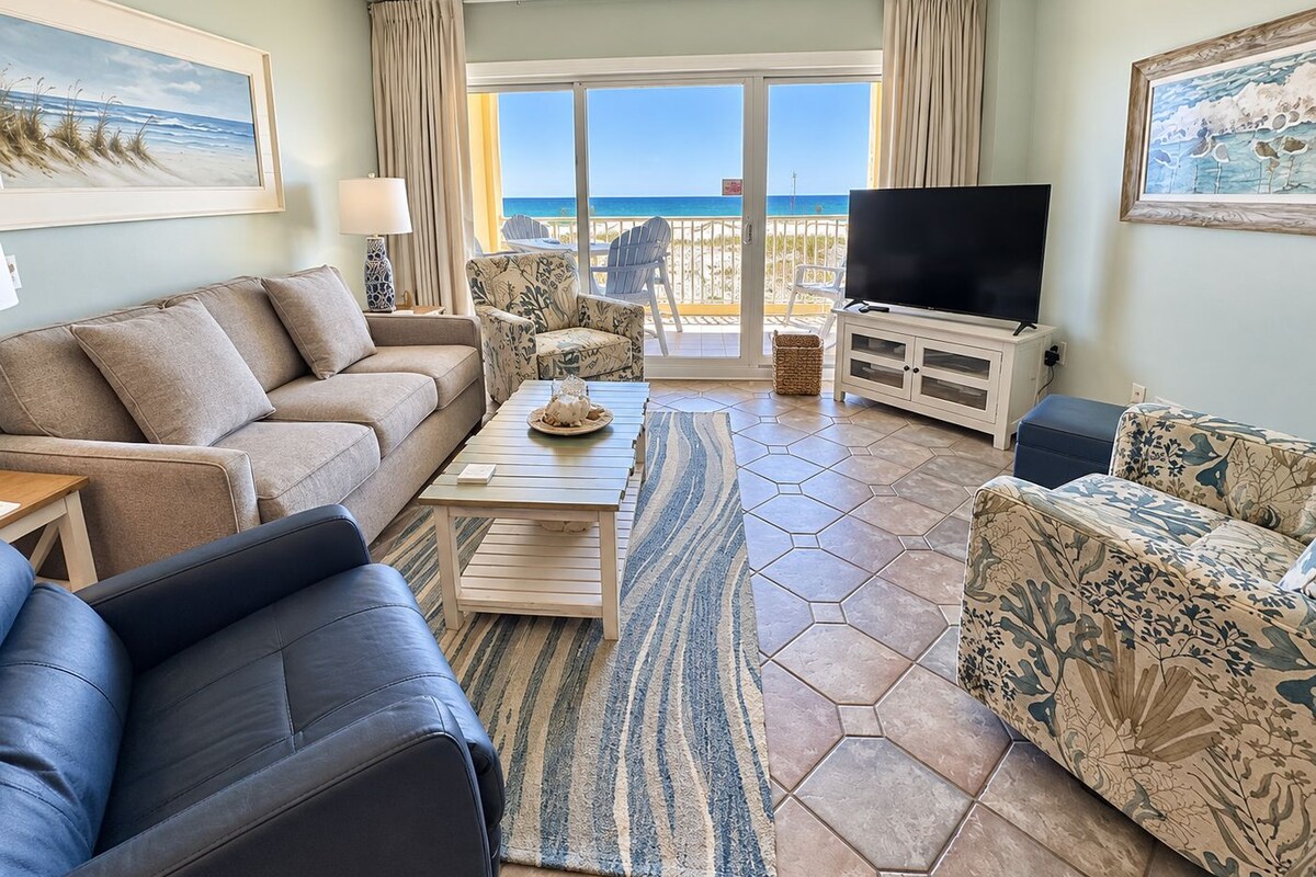 Ein Wohnzimmer mit einer Couch, Sesseln, einem Couchtisch und einem Flachbildfernseher. Es gibt einen Balkon mit Meerblick.