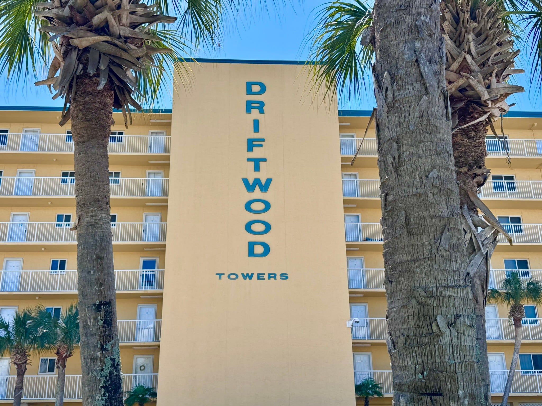 Egy épület, amelynek homlokzatán a „DRIFTWOOD TOWERS” felirat látható, és melyet pálmafák öveznek.