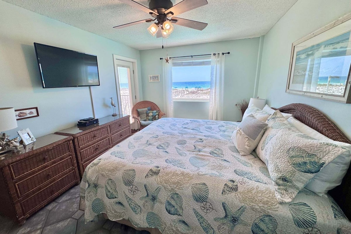 A bedroom with a bed, dresser, TV, ceiling fan, and a view of the beach.
