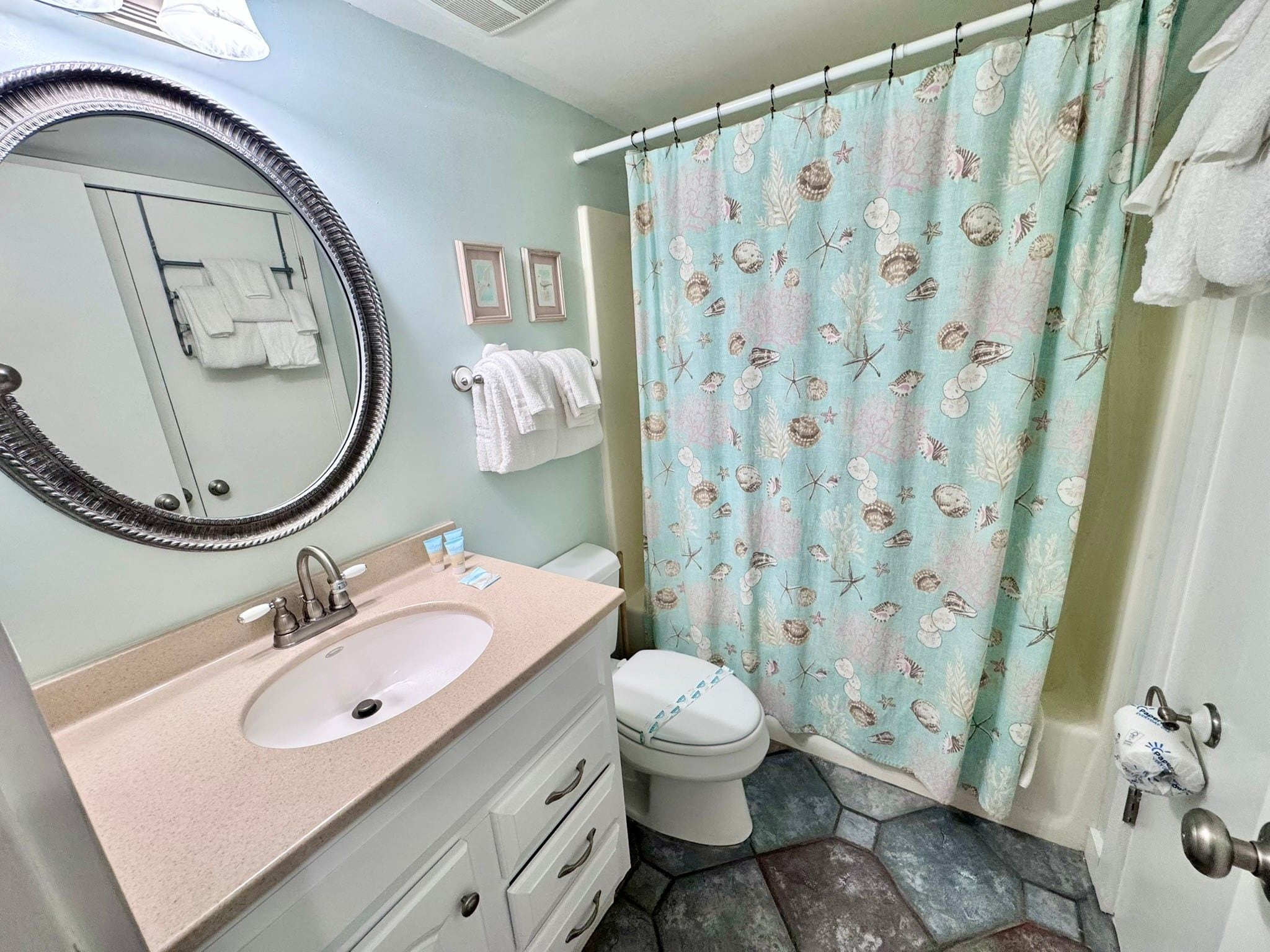 A bathroom with a sink, mirror, toilet, and a shower curtain with a sea shell pattern.
