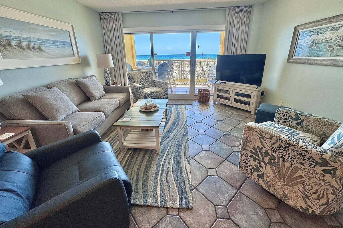 Una sala de estar con sofá, sillón, mesa de centro y televisor, con vista al mar a través de un ventanal.