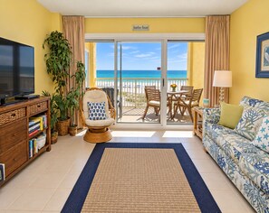 Una sala de estar con televisor, sofá, sillón, mesa y vista a la playa a través de una puerta corrediza.