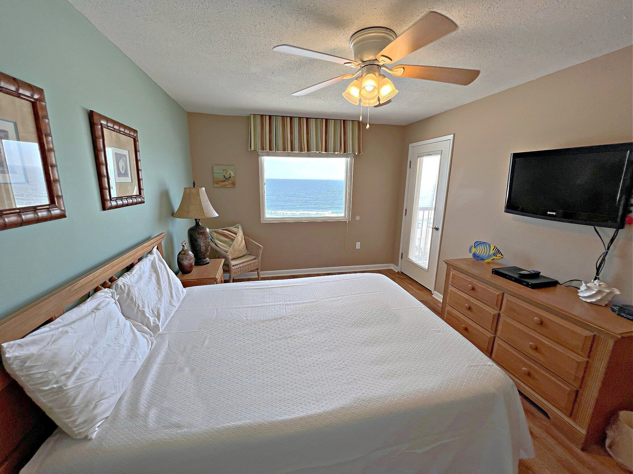 A hotel room with a bed, a ceiling fan, a television, and a window with a view of the ocean.