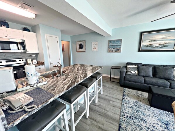 A modern kitchen with a marble countertop, white cabinets, and a built-in oven. There is a dining area with a black leather couch and a blue patterned rug. - Grand Beach Resort (Gulf Shores)