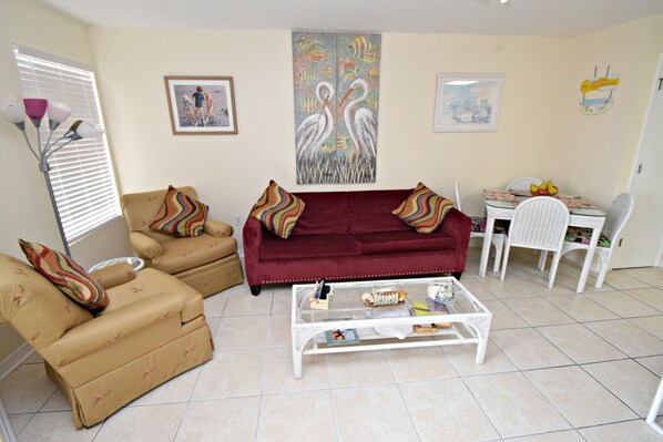 A living room with a red sofa, beige armchairs, and a white coffee table. There are two framed pictures on the wall and a dining area with a table and chairs. - Grand Beach Resort (Gulf Shores)