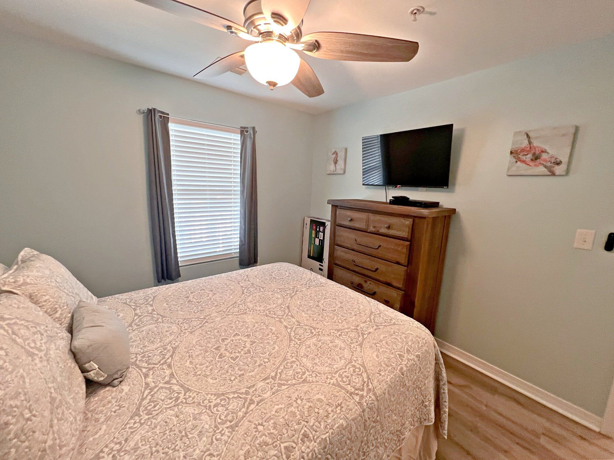 A bedroom with a bed, a ceiling fan, a dresser, a television, and a window with curtains.