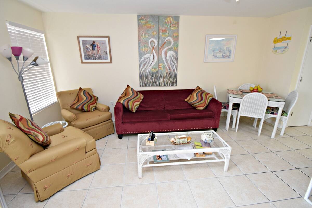 Een woonkamer met een rode bank, beige fauteuils en een witte salontafel. Twee ingelijste foto's hangen aan de muur en er is een eethoek met een tafel en stoelen.