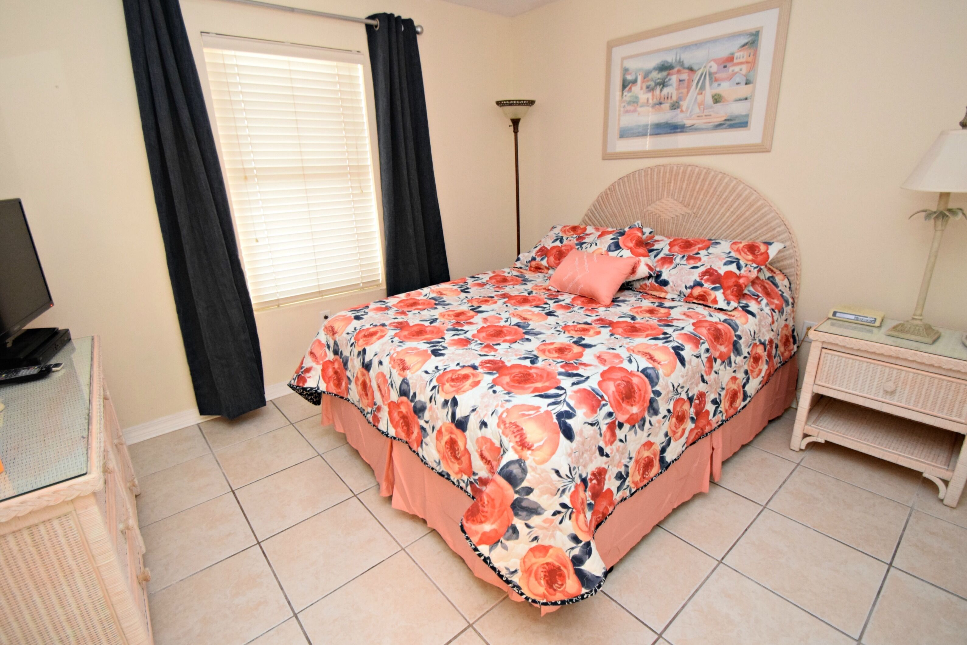 A bedroom with a floral bedspread, a bedside table, a lamp, and a painting on the wall.