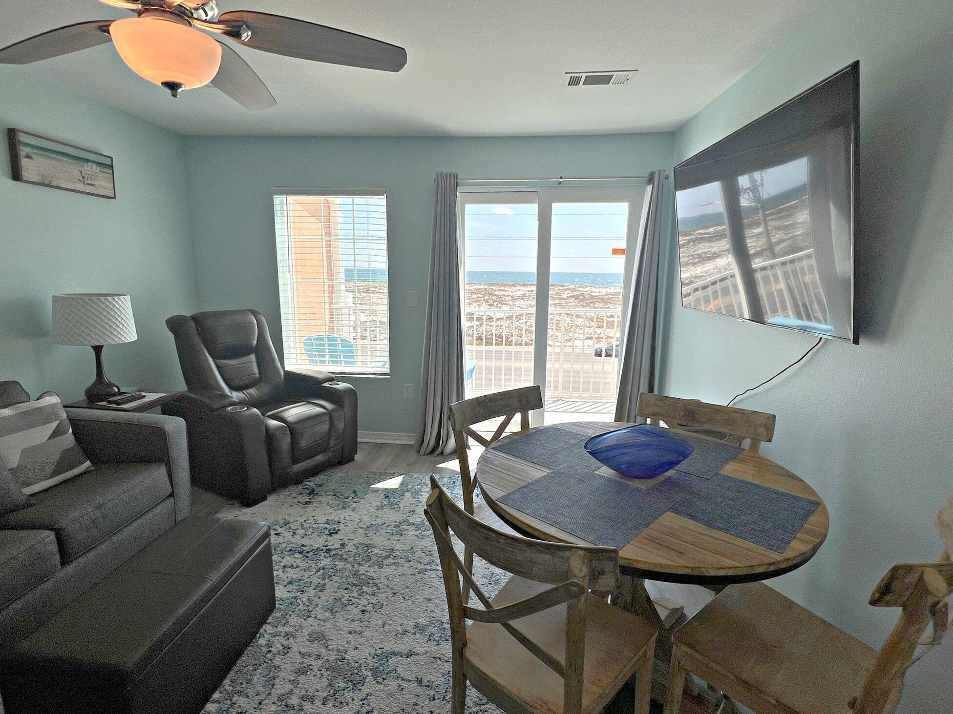 A living room with a sofa, a recliner, a dining table with chairs, a ceiling fan, a TV mounted on the wall, and a view of the beach through the windows.