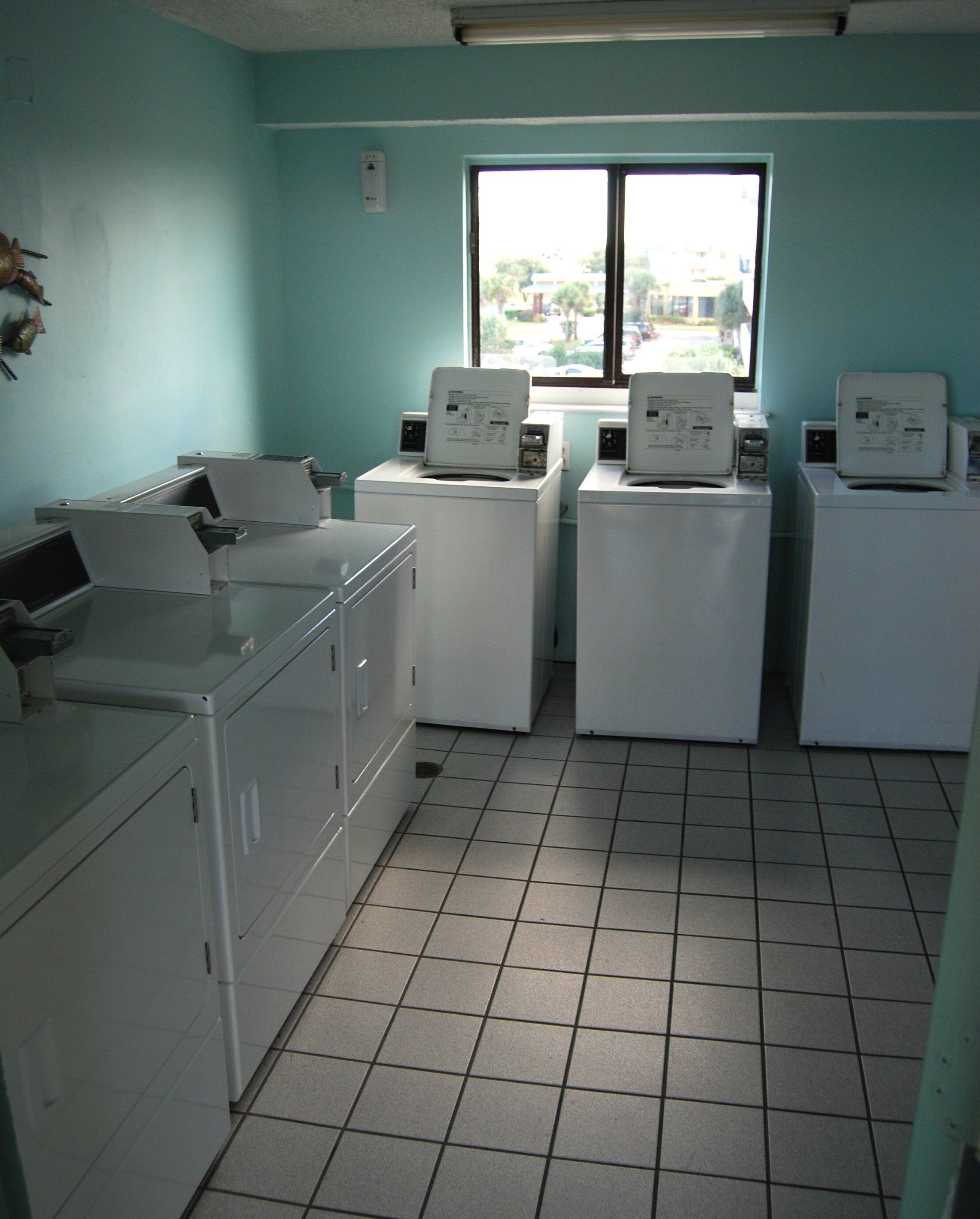 A laundry room with four washing machines and a dryer, a window with a view of buildings outside, and a tiled floor.
