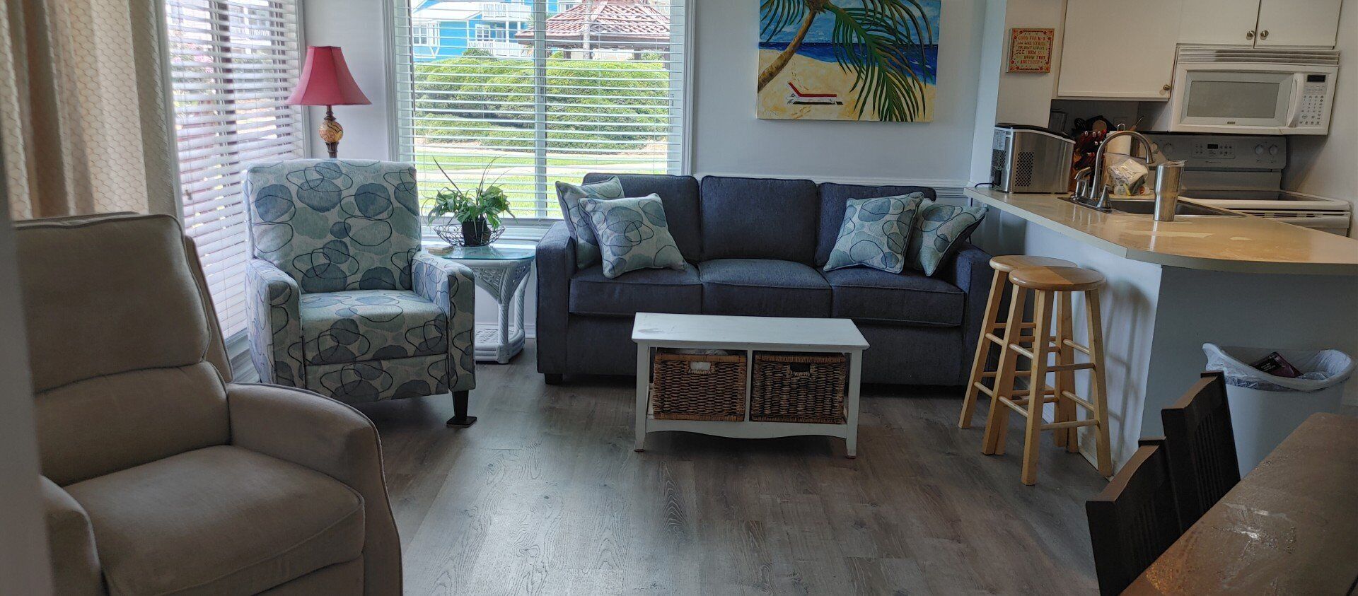 A living room with a sofa, armchair, coffee table, and a small side table with a lamp. There is a kitchen area with a microwave and a sink.