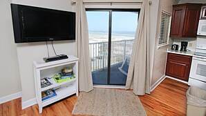 Uma sala de estar com televisão de tela plana, um móvel branco para entretenimento com livros, um tapete estampado e vista para a praia através de uma porta de vidro deslizante.