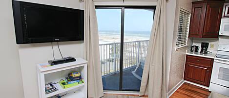 Una sala de estar con televisor de pantalla plana, un mueble blanco para equipo audiovisual con libros, un tapete estampado y vista a la playa a través de un ventanal corredero.