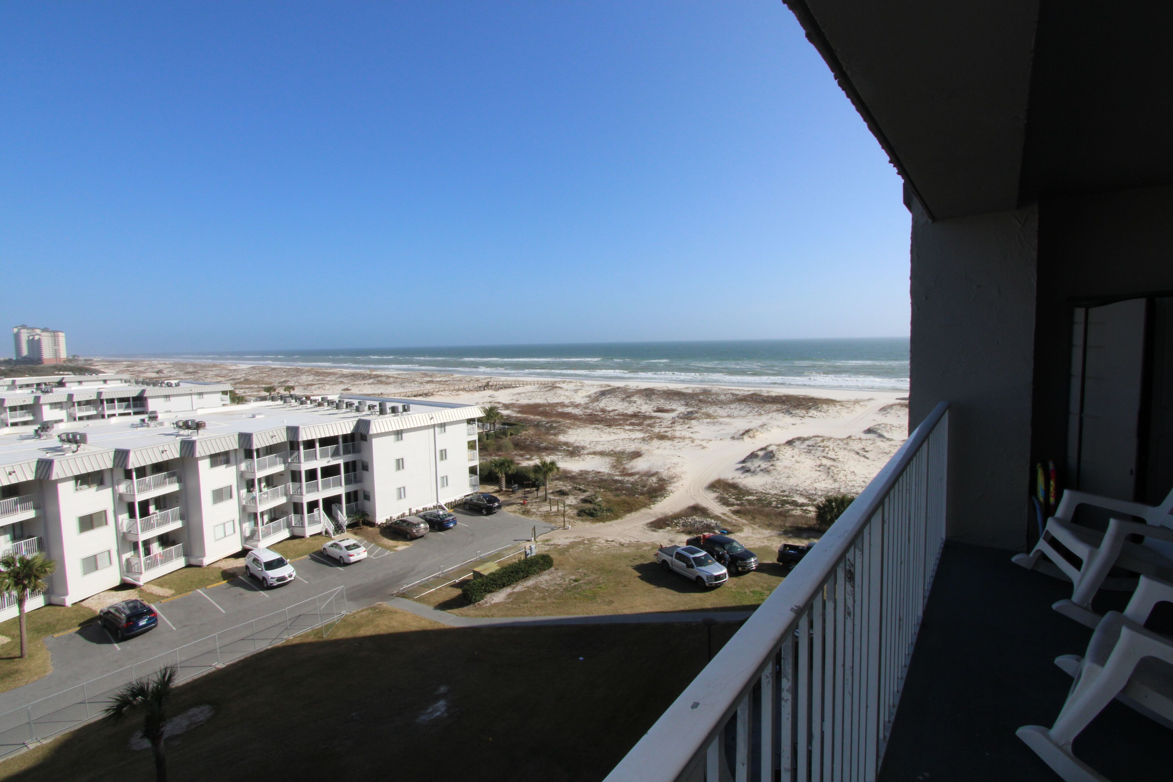 A view from a balcony overlooking a beach with a parking lot and a multi-story building with balconies.