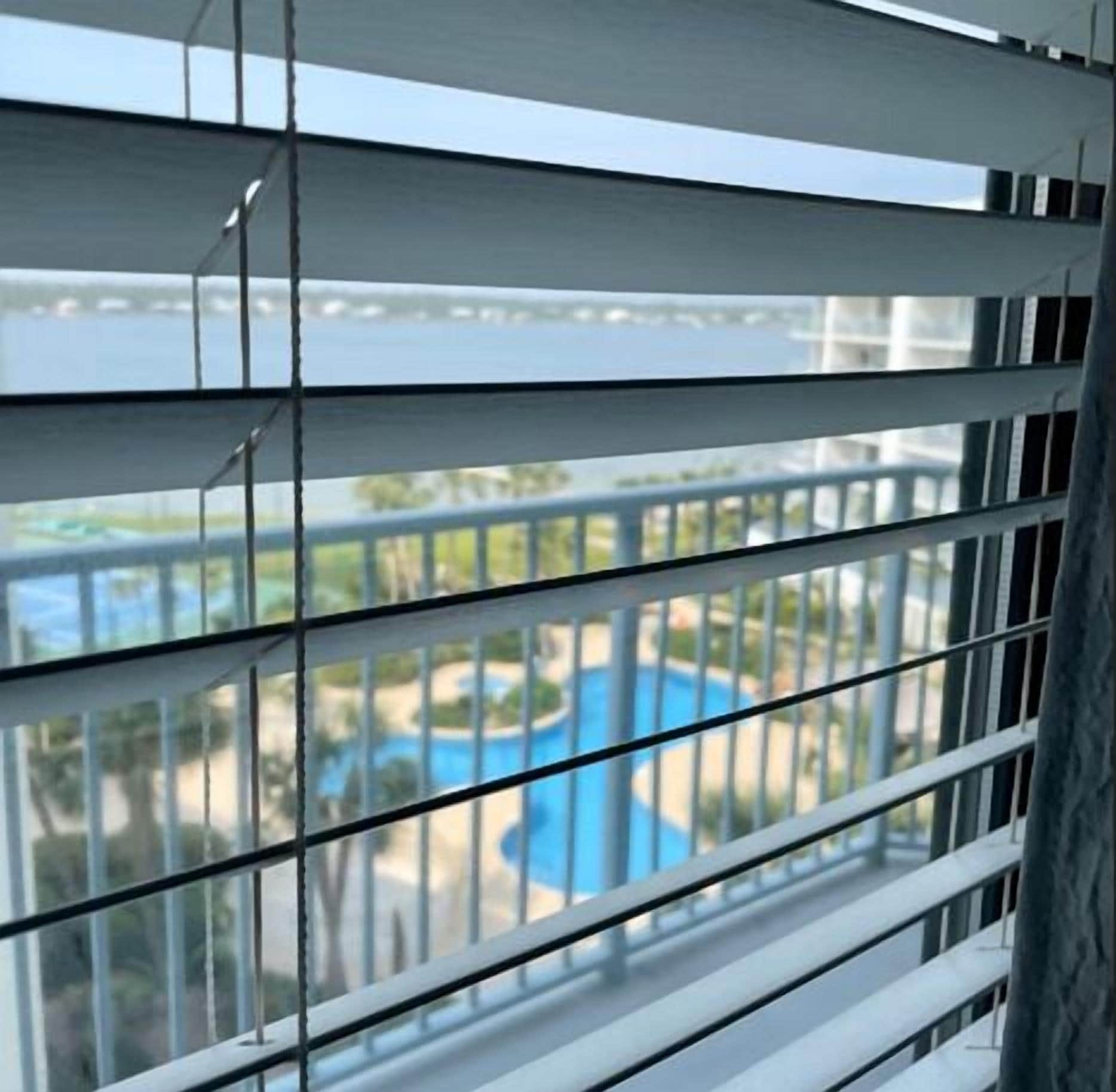A view through window blinds of a pool area with a clear sky and a body of water in the background.