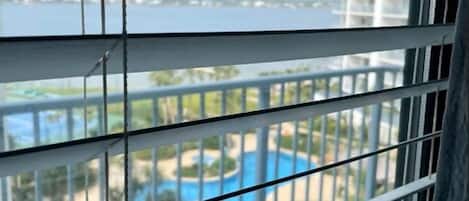 A view through window blinds of a pool area with a clear sky and a body of water in the background.
