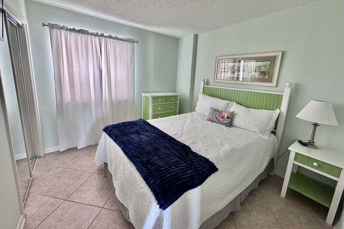 A bedroom with a bed, a nightstand, a dresser, a window with curtains, and a framed picture on the wall.