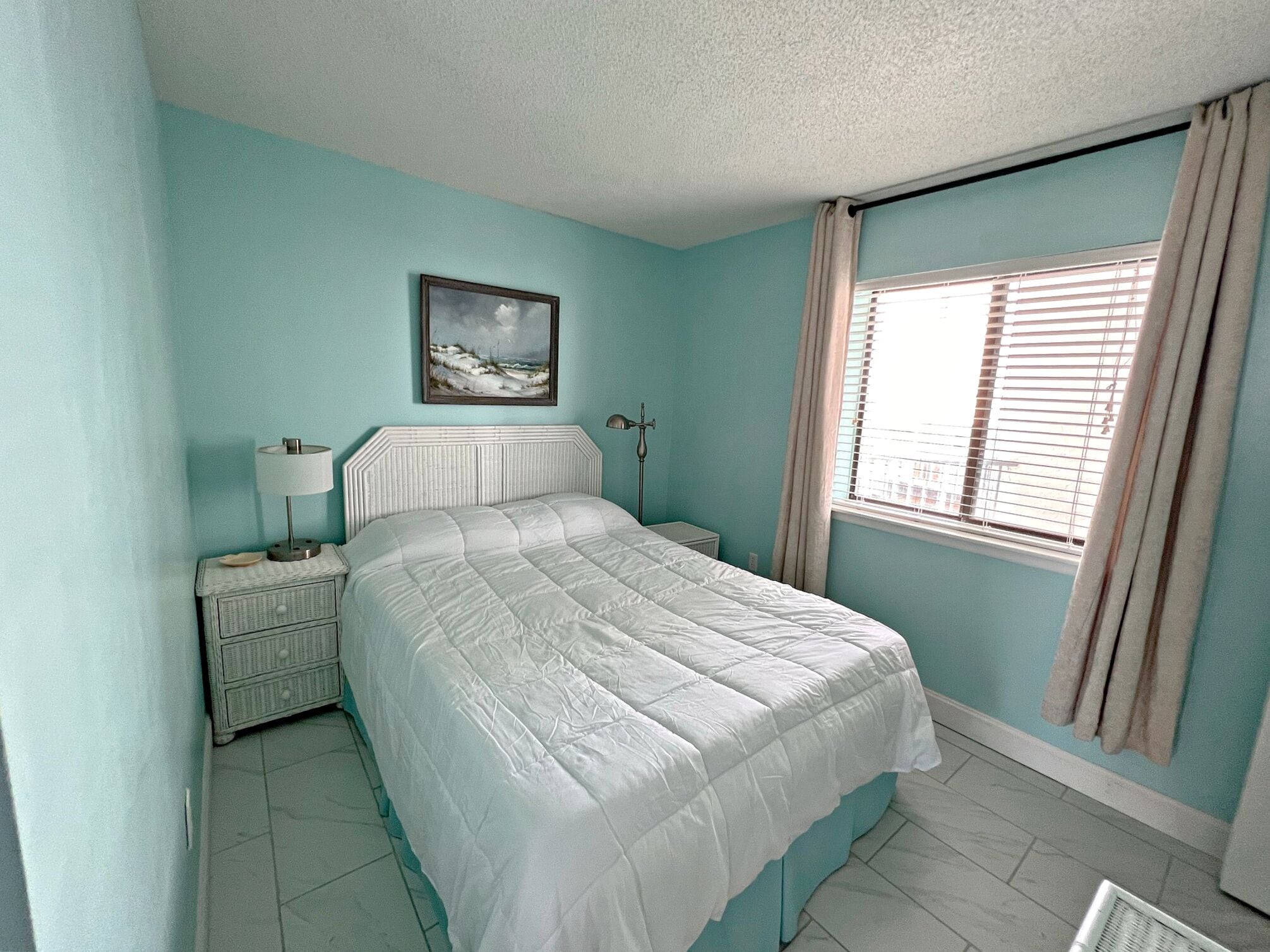 A bedroom with a bed, a nightstand, a lamp, a picture on the wall, and a window with curtains.