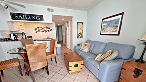 A living room with a sofa, chairs, a coffee table, a dining area with a table and chairs, a kitchen area, and a sign that reads "SAILING". - Gulf Shores Surf & Racquet Club (Gulf Shores)
