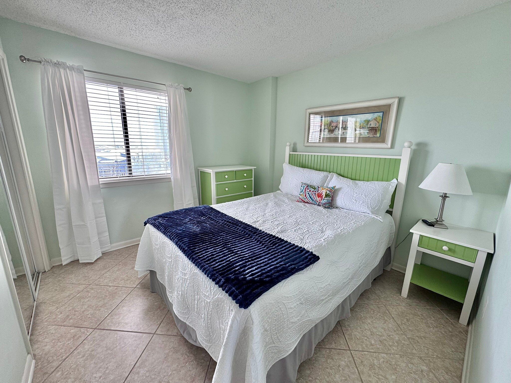 A bedroom with a bed, a nightstand, a lamp, a window with curtains, and a green dresser.