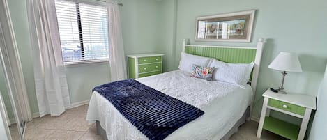 A bedroom with a bed, a nightstand, a lamp, a window with curtains, and a green dresser.