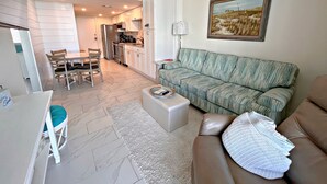 A modern living room with a sofa, a dining table with chairs, a kitchen area, and a painting on the wall. - Gulf Shores Surf & Racquet Club (Gulf Shores)