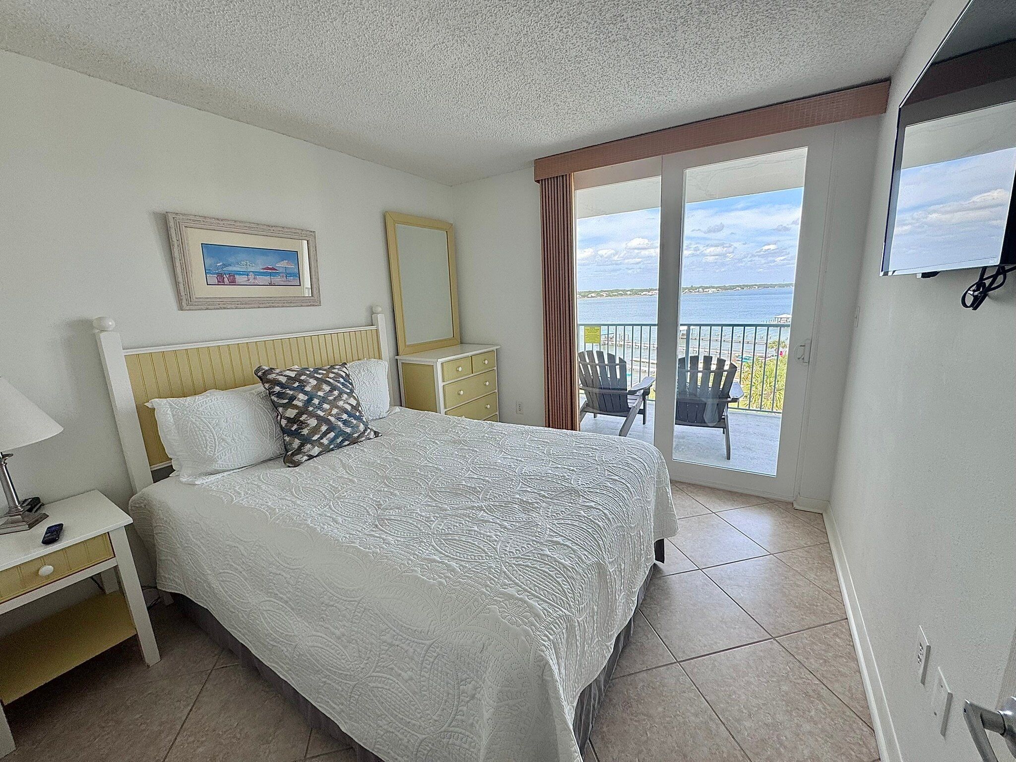 A hotel room with a bed, a nightstand, a dresser, a large window with a view of the sea, and a mounted television.