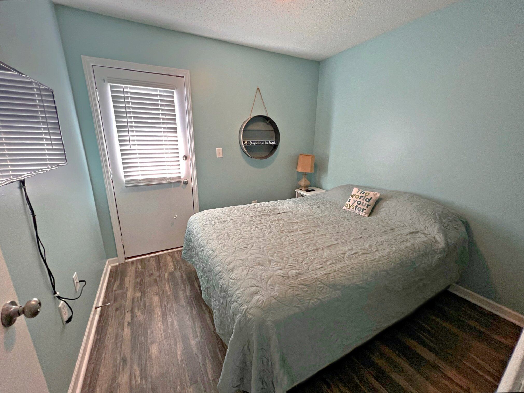 Quarto com cama, porta, luminária na parede e pequena mesa com abajur.