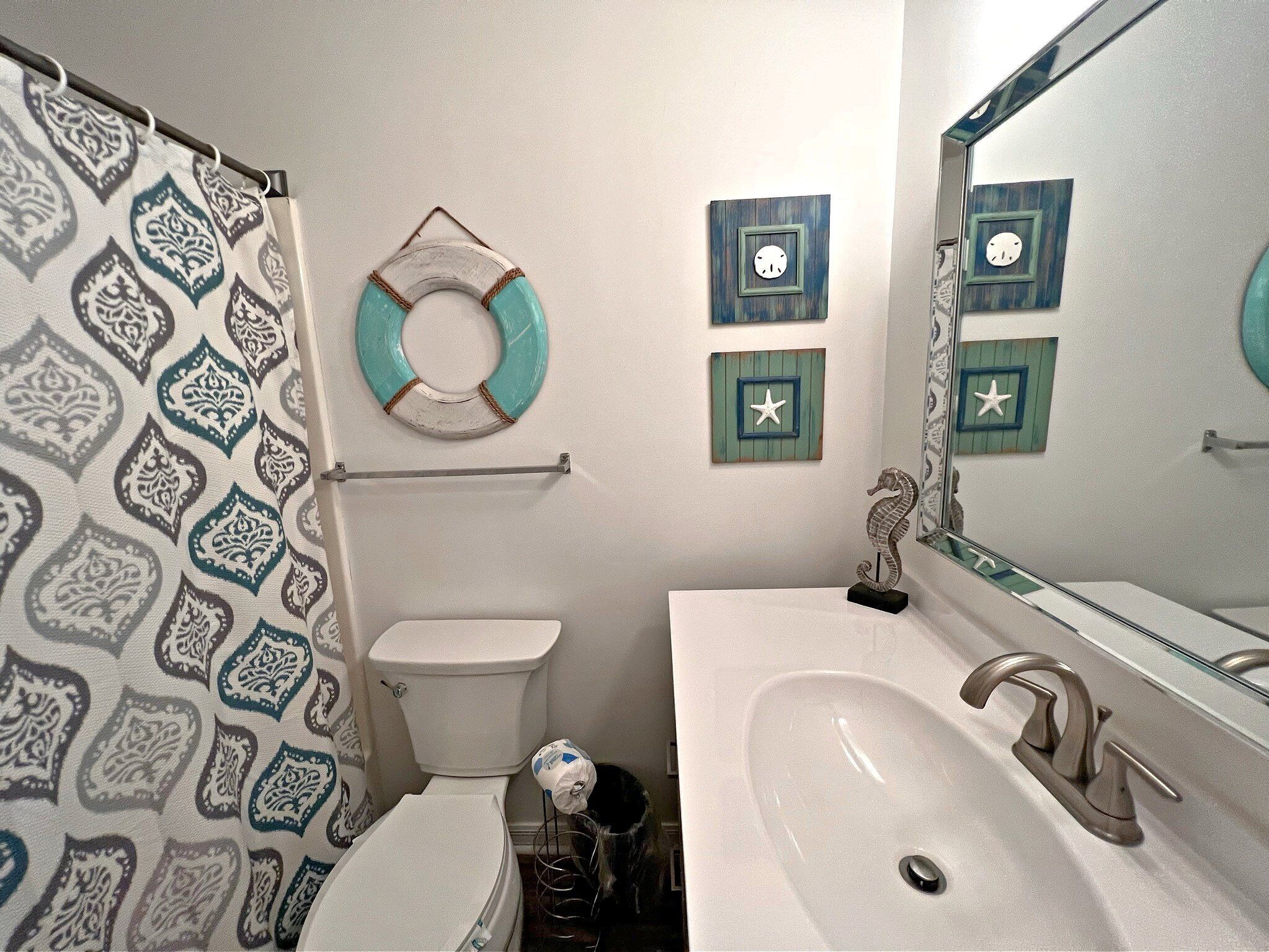 Un baño con cortina de ducha estampada, un inodoro, un lavamanos con espejo y detalles decorativos en la pared, incluyendo un caballito de mar y una estrella de mar.