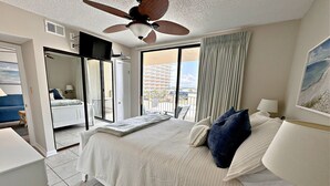 A hotel room with a bed, a ceiling fan, a TV, a balcony with curtains, and a painting on the wall. - Gulf Village (Gulf Shores)