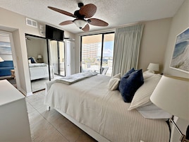 A hotel room with a bed, a ceiling fan, a TV, a balcony with curtains, and a painting on the wall.