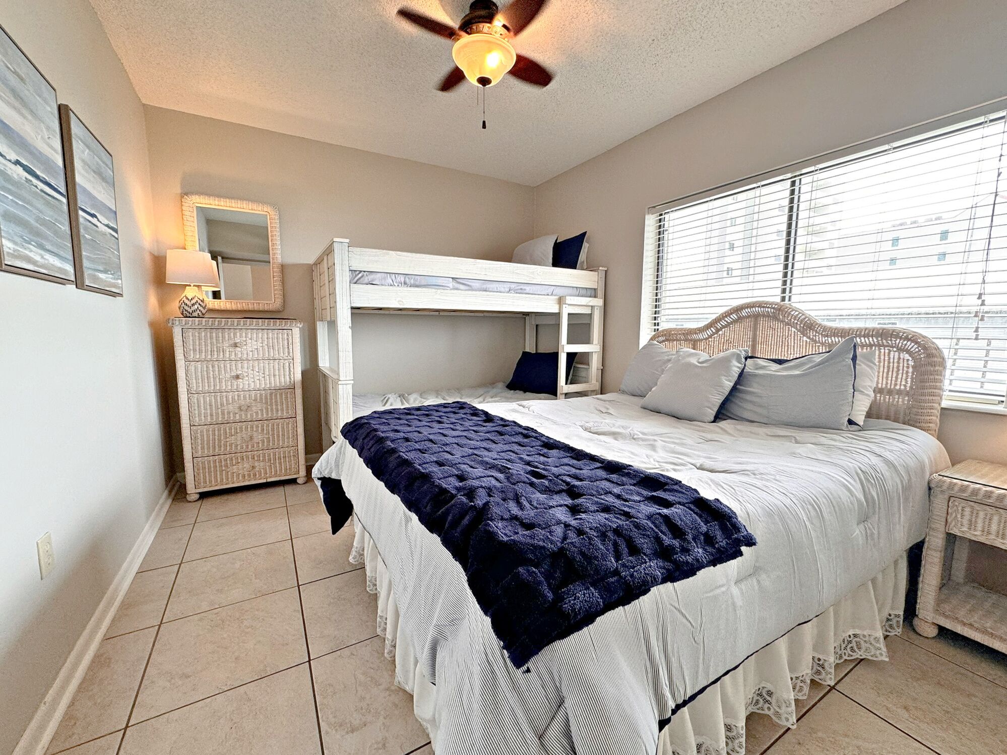 A bedroom with a bunk bed, a bedside table, a lamp, a dresser, and a window with blinds.