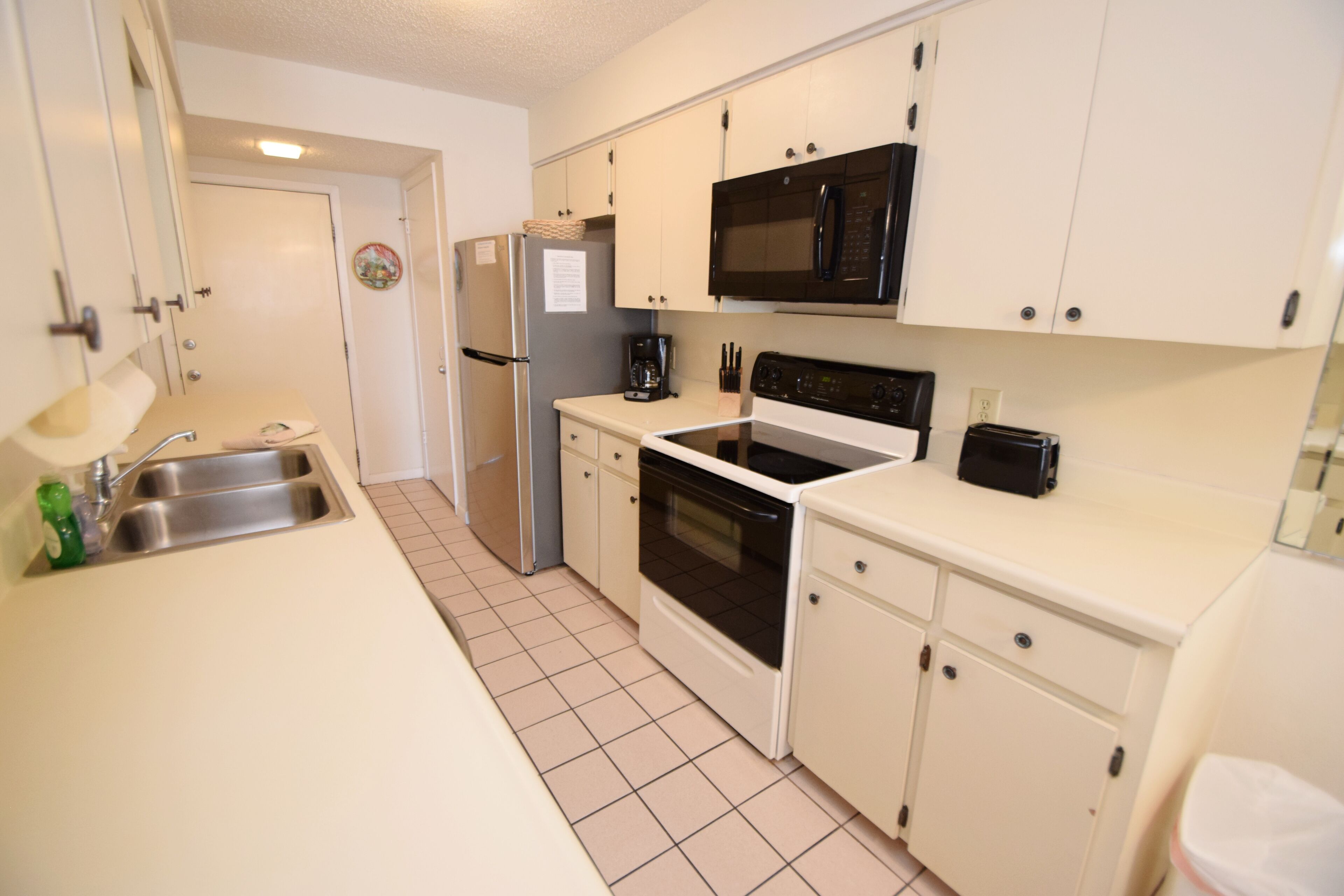 A compact kitchen with white cabinets, a stainless steel sink, a black microwave, a stove, and a toaster.
