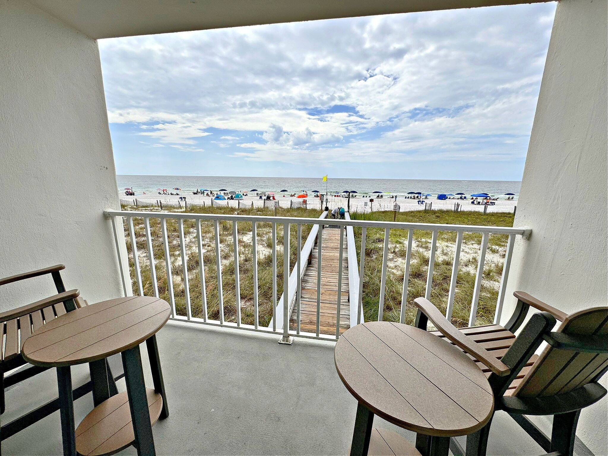 A balcony with a view of the beach, featuring a wooden deck, white railing, and two wooden tables with chairs.