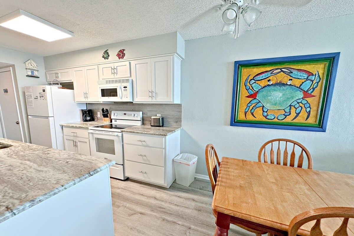 A compact kitchen with white cabinets, a microwave, a stove, and a sink. There's a wooden dining table with chairs and a colorful crab painting on the wall.
