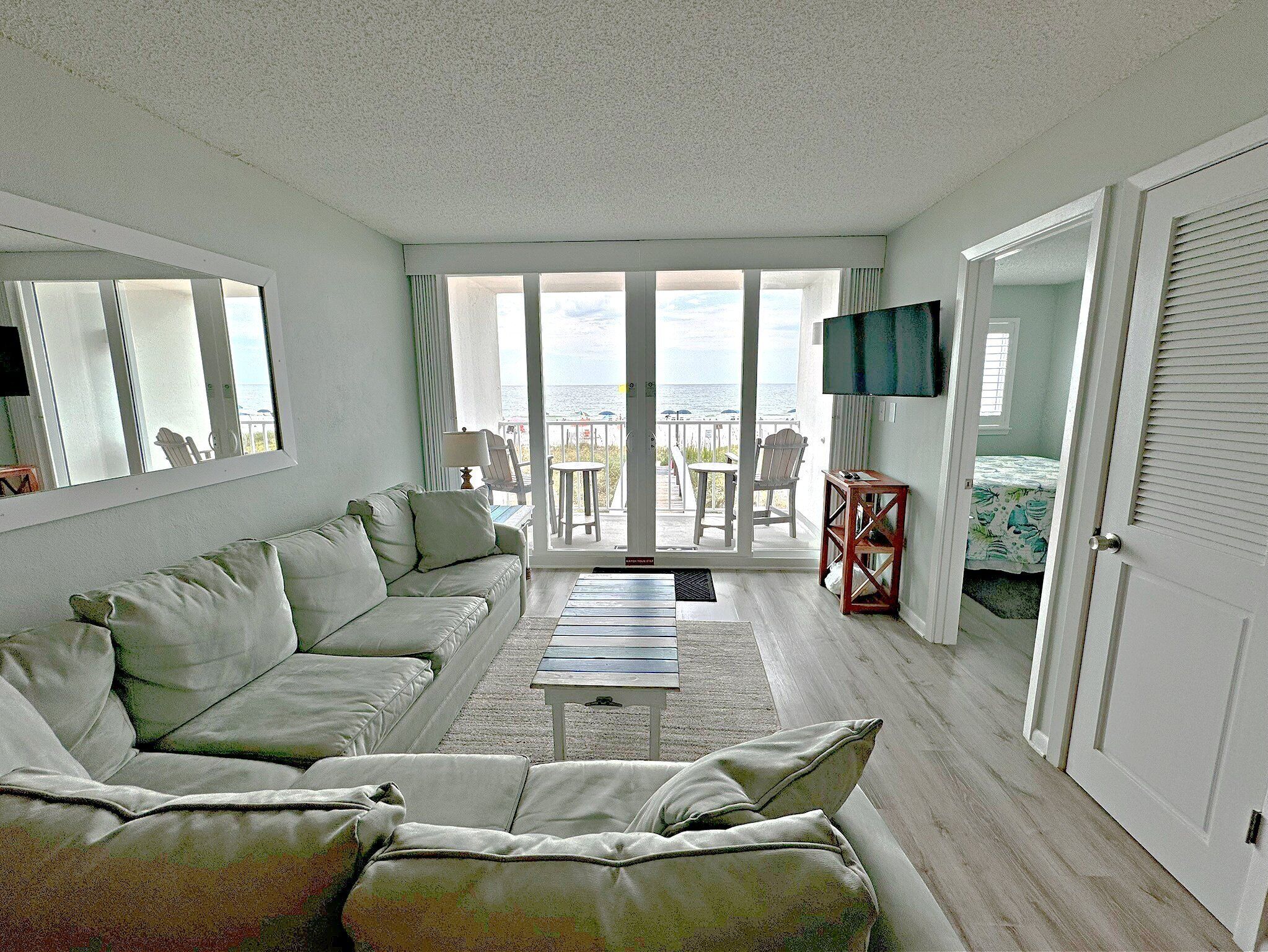 A living room with a sofa, a coffee table, and a TV. There is a balcony with a view of the ocean.